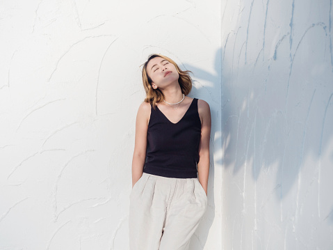 Asian woman look cool with short hair wearing black tank top and white trousers standing and keep hands in pockets on white wall background, close eyes. Female summer fashion minimal style.