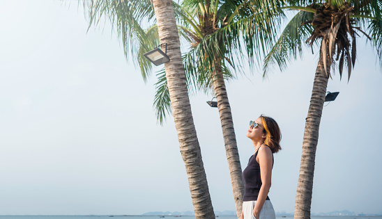 Asian woman look cool with short hair wearing sunglasses, black tank top and white trousers standing and keep hands in pockets on the beach under the coconut palm trees and sea background, summertime.