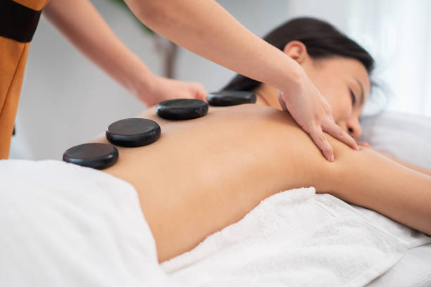 ウェルネスセンターのセラピールームでスパホットストーンマッサージを受ける美しい女性。マッサージ師または理学療法士が肩と首でマッサージをしています。女の子はボディトリートメ� - massaging spa treatment stone massage therapist ストックフォトと画像