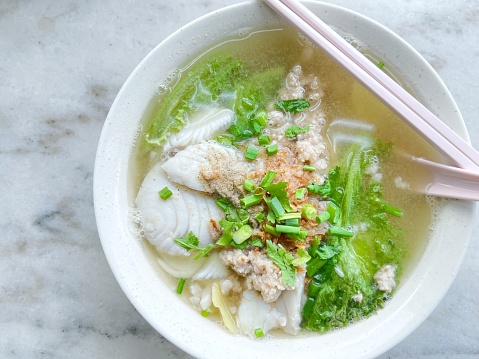 Overhead view of a bowl of freshly served Teochew fish slice soup with rice vermicelli and greens