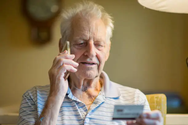 A senior man is making a telephone call with his smart phone, he appears to be reading the information off of a credit or debit card while on the phone. He is sitting down in an armchair at home.