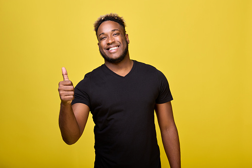 A satisfied young man shows a positive hand gesture, he is satisfied with everything. A dark-skinned, cheerful young man points his thumbs up while standing on a yellow background.