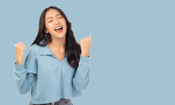 jovem asiática animada com as mãos levantadas celebrando o sucesso retrato alegre mulher asiática positiva com as mãos levantadas e punhos de boca aberta menina ficar feliz, desfrutar da vida isolada no espaço de cópia azul - arms raised behavior isolated one person - fotografias e filmes do acervo