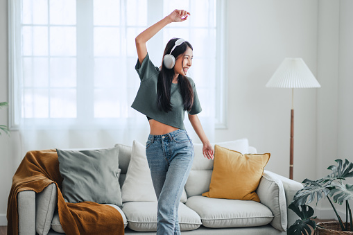 Happy carefree asian woman wearing headphones have fun moving listening to music relax in living room, She dance enjoy leisure weekend at home.