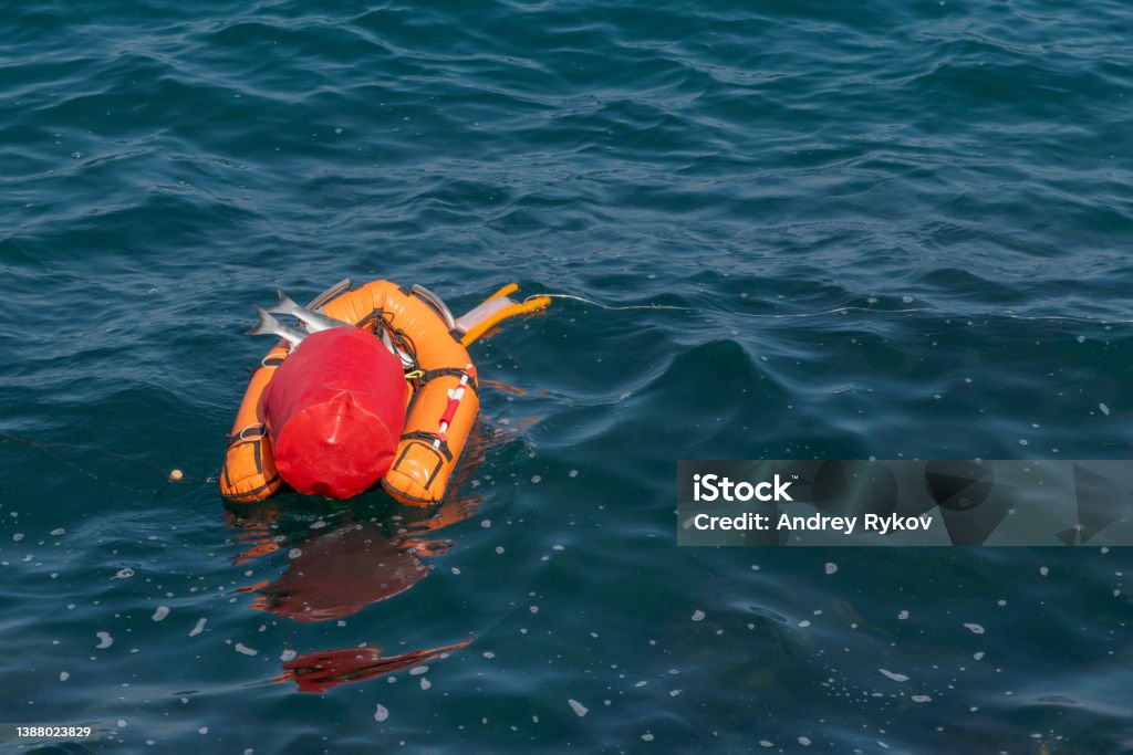 Orange buoy-boat with fishing tackle Orange buoy-boat with fishing lures in form of fish for spearfishing and red bag floats on surface of blue sea water. Copy space for your text. Hobbies and leisure theme. Bag Stock Photo