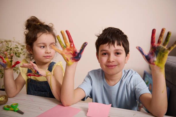 Two adorable European children , boy and girl sitting close to each other and showing their stained hands in colorful watercolor paints or gouaches, cutely smiling looking at camera, during art class Adorable European children, brother and sister sitting close to each other and showing their stained hands in colorful watercolor paints or gouaches, cutely smiling looking at camera, during art class cutely stock pictures, royalty-free photos & images