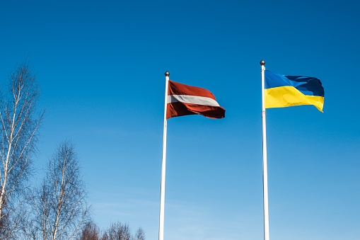 Against the backdrop of a blue sky, the Ukrainian and Latvian flags hang on two flagpoles. Symbol of support for Ukraine from Latvia and Europe.
