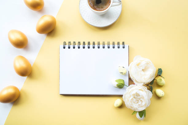 istock Golden easter eggs, coffee mug, delicate flowers and notebook with place for text on on yellow background. Trendy flat lay. minimal concept. 1388052491
