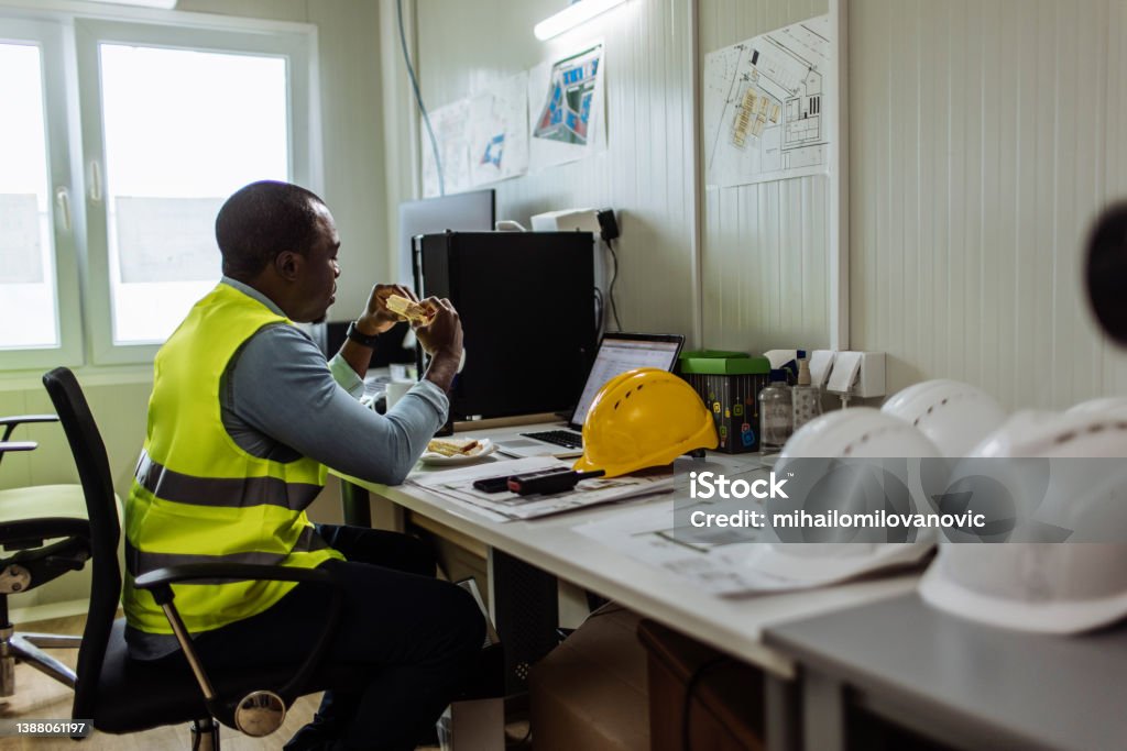 Lunch break in the office African American man eating a sandwich while working in his office at the construction site Construction Site Stock Photo
