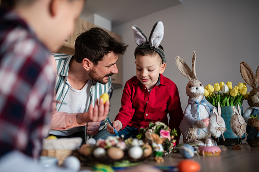 Happy young family with two kids celebrating Easter at home. They are smiling, painting Easter eggs and wearing bunny ears. Happy Easter. Easter 2022.