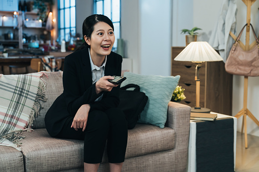portrait asian career woman leaning forward and holding remote control is binge-watching after work. taiwanese female in black suit is showing great interest in the plot development of the tv series