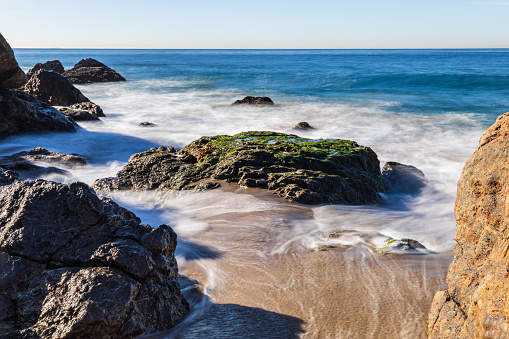 Rocky beach at Malibu, California. Waves receding from the sandy shore; blue Pacific Ocean in the background.