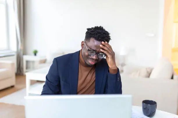 Stressed-out young businessman sitting with his head in his hands while working at a desk in an office. Portrait of frustrated young man with laptop working indoors, home office concept.