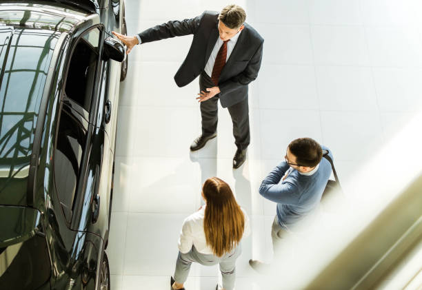 Above view of happy salesman talking to young couple in car showroom. High angle view of happy mid adult salesperson talking to young couple in a car showroom. Copy space. car dealership stock pictures, royalty-free photos & images