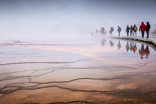 Wyoming, USA - October 1, 2019: Misty scene of walking tourists on wooden boardwalk inside orange wet surface of Grand Prismatic Spring, the most famous place in Yellowstone national park.