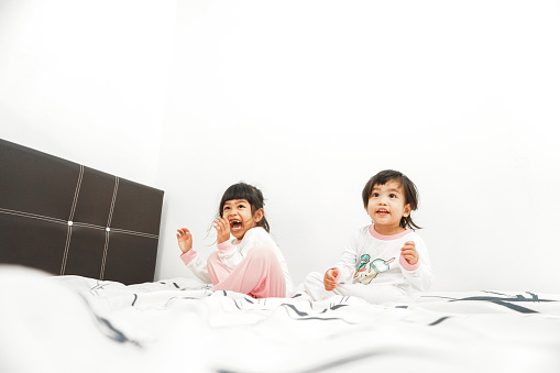 Happy kids playing in white bedroom. Little girls, sisters play on the bed wearing pajamas. Family at home