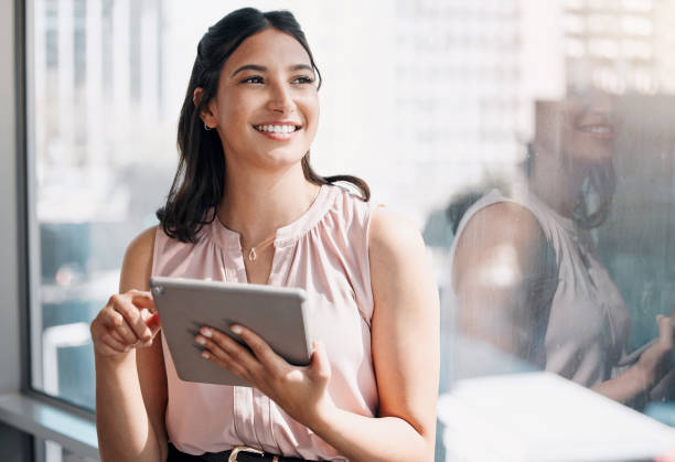 Shot of an attractive young businesswoman standing alone in the office and looking contemplative while using a digital tablet My goals are in clear sight smiling businesswoman stock pictures, royalty-free photos & images