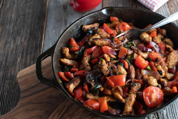 Healthy pan dish with fried meat strips and vegetables such as red onions, leek, tomatoes, garlic and bell peppers. Served in a cast iron skillet on wooden table. Closeup view