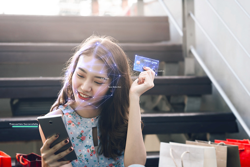 Facial scan confirm before online transaction concept. Young adult asian woman using credit card for shopping and pay with digital wallet via mobile phone.