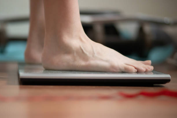 piedi sulla bilancia, una donna viene pesata prima di fare sport, un bilanciere e manubri sullo sfondo - weight scale dieting weight loss foto e immagini stock
