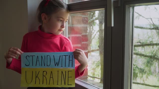 Girl looking outside window and holding banner with inscription Stand with Ukraine at blue yellow flag
