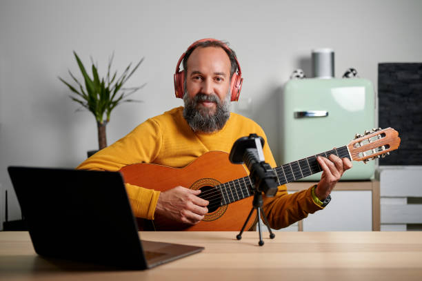 istock Bearded mature man in yellow sweatshirt and wireless headphones looking at camera while playing guitar and recording audio for blog at home 1388134710