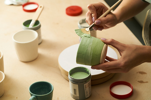 Primer plano de la chica pintando una taza de arcilla con esmalte. Mujer coloreando cerámica en taller con un pincel. Pintor en delantal verde acristalamiento de olla de barro photo