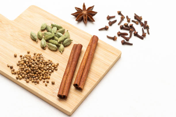 Coriander seeds, cinnamon sticks, and cardamom on wooden board. Cloves and star anise on table. Top view. White background