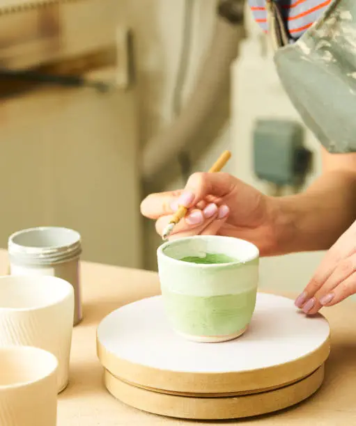 Close-up of girl painting clay mug with glaze. Woman coloring pottery in workshop with a paintbrush. Painter in green apron glazing clay pot.