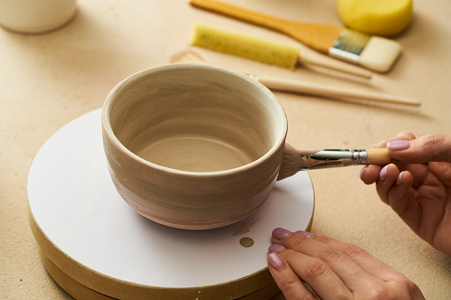 Close-up of girl painting clay mug with glaze. Woman coloring pottery in workshop with a paintbrush. Painter in green apron glazing clay pot.