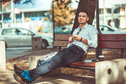 Attractive young man is sitting alone on bench in park, looking peacefully with coffee and book near him