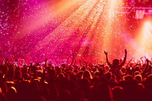 A crowded concert hall with scene stage in red lights, rock show performance, with people silhouette, colourful confetti explosion fired on dance floor during a concert festival