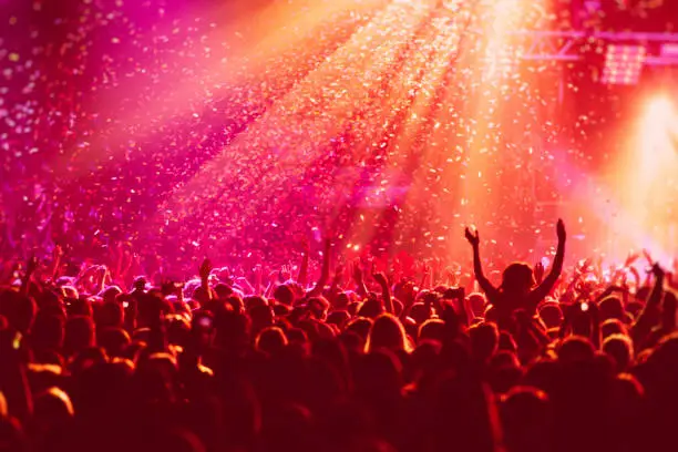 Photo of A crowded concert hall with scene stage in red lights, rock show performance, with people silhouette, colourful confetti explosion fired on dance floor air during a concert festival