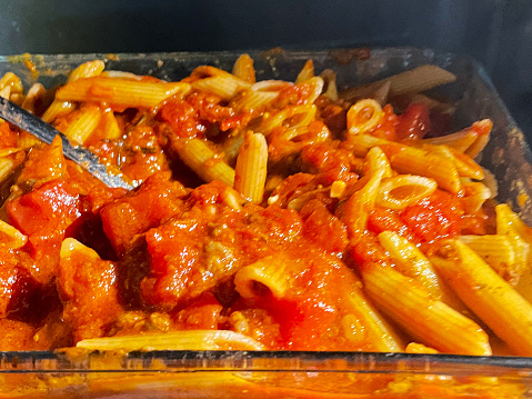 Stock photo showing close-up, elevated view of glass, oven dish containing penne pasta covered in rich tomato sauce in microwave oven being stirred by a metal spoon.
