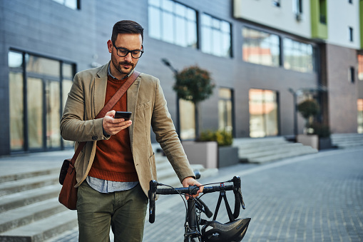 Fashionable, cheerful young businessman walking through town. He is pushing his bike along a pedestrian walkway, text messaging from his phone