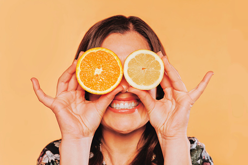 Close up portrait of happy smiling brunette woman holding half an orange and half lemon covering her eyes, isolated on yellow background. Summer citric concept.