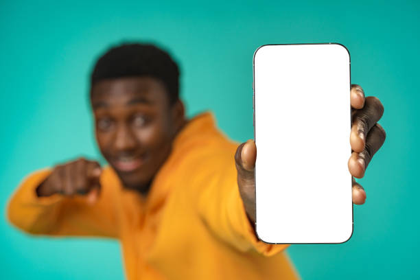 istock Smiling casual black man pointing at blank white cell phone screen in studio 1388169209