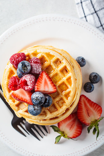 Homemade waffles with berries, powdered sugar and maple syrup on a white plate, gray background, top view.