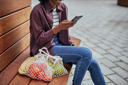 Young african woman sitting on bench outside after shopping and using cellphone