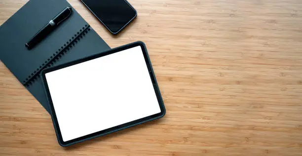 Top view of blank white screen digital tablet, notebook, pen and mobile phone on wooden table, copy space.