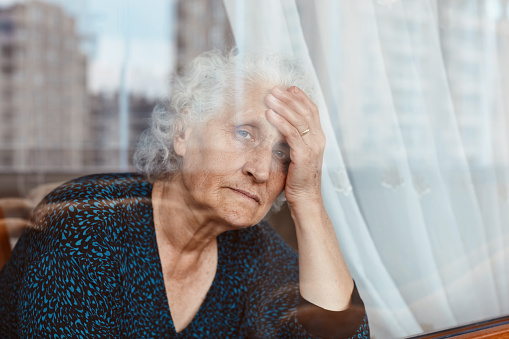 Senior woman sitting alone and looking at camera out the window lost in deep thoughts and sadness