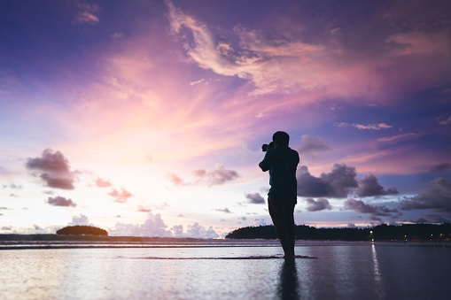 Silhouette rear view of adult travel asian photographer man with camera on beach sand with beautiful dramatic sunset sky. Outdoor domestic tourism at andaman ocean. Phuket, Thailand.