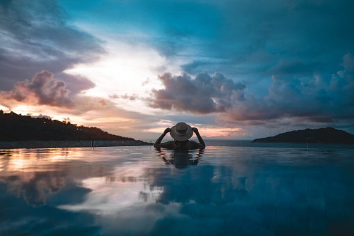 Summer travel on holiday at southeast tropical concept. Rear view asia woman relax at outdoor pool resort sea panorama view. With straw hat and dramatic sunset sky. Phuket island, Thailand