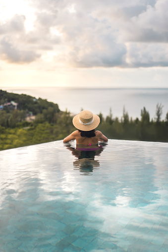 Summer travel on holiday at southeast tropical concept. Rear view asia woman relax at outdoor pool resort sea panorama view. With straw hat and dramatic sky. Phuket island, Thailand