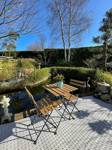 Stock photo showing ornamental Japanese-style garden with outdoor lounge area in Spring. Featuring a large expanse of white, interconnecting, white plastic decking tiles with outdoor patterned rug, providing a family space for outdoor hardwood seating.