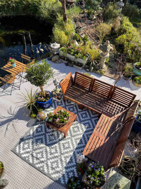 Stock photo showing ornamental Japanese-style garden with outdoor lounge area in Spring. Featuring a large expanse of white, interconnecting, white plastic decking tiles with outdoor patterned rug, providing a family space for outdoor hardwood seating. imagen de área de descanso al aire libre en primavera, asientos de madera dura, mesa de madera con centro de mesa de plantas con flores, bonsáis, arces japoneses, estanque koi, jardín de diseño oriental ajardinado, día soleado, vista elevada