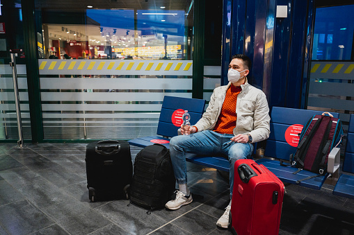 Young man with protective face mask sitting in a chair in Bologna airport in Italy by night. Guy holds a bottle of water and he has with many baggage.