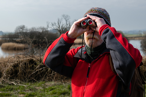 Close up front view of a senior man standing next to a river in a remote rural location using binoculars