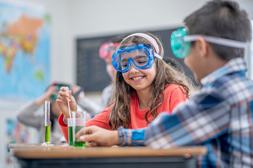 A group of elementary students sit side-by-side at a long table, as they each explore different aspects of science class. The focus is on a girl in a bright orange shirt who is holding a pipette as she prepares to draw out some of the green liquid in the beaker in front of her. She has protective goggles on her face and is smiling. Her peers can be seen working with other experiments on either side of her.