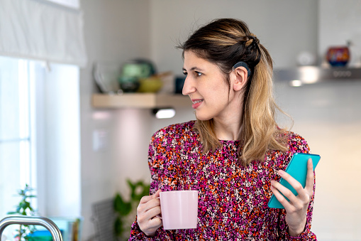 A visually impaired young woman  with hearing aid, standing in the kitchen  and using  a smartphone. She does not wear eyeglasses.